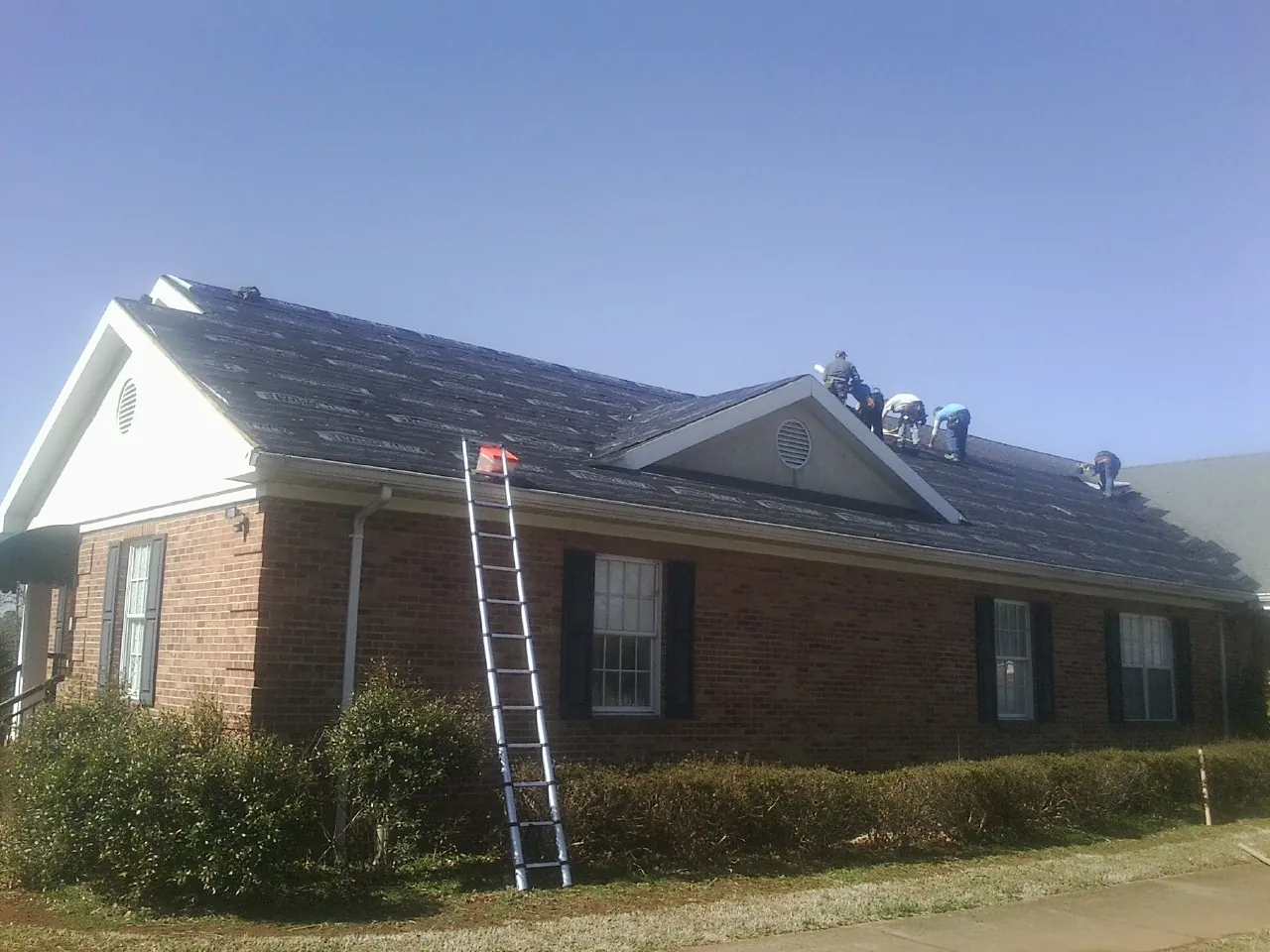 Crew tearing shingles off a brick ranch roof with a ladder set — Metal Roof Repair work by our crew in Nielsville