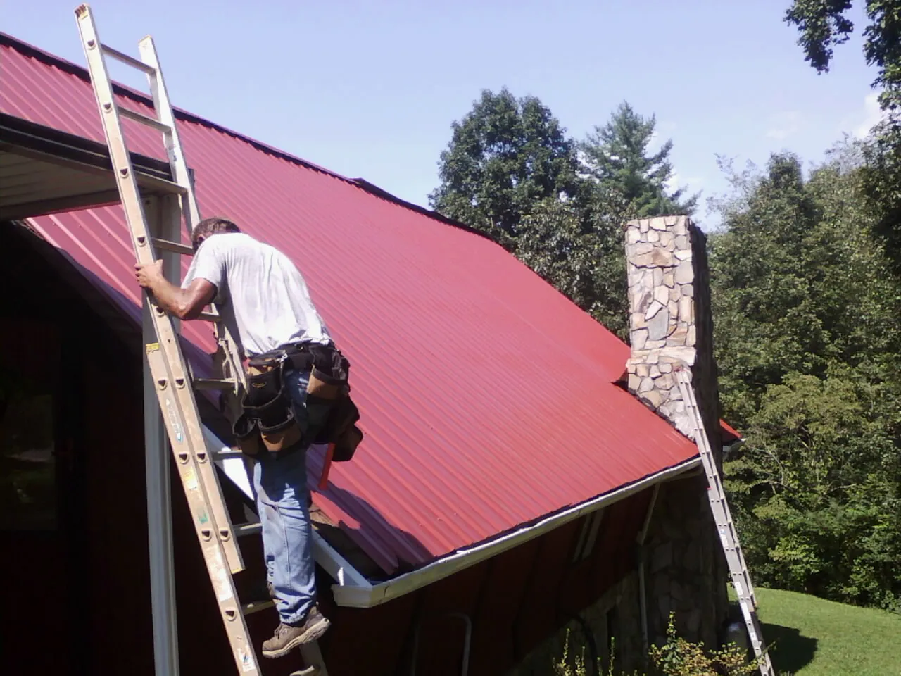Roofer on a ladder beside a red metal roof and stone chimney — Soffit & Fascia Repair by Roofing Brothers in Nielsville
